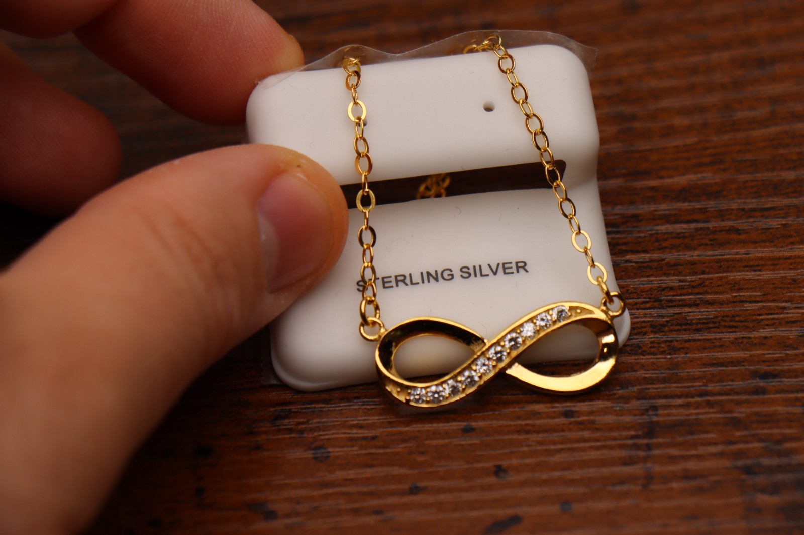 A hand holds a 14k gold over 925 Sterling Silver Infinity Necklace with a CZ pendant on an 18" chain, displayed on a white card labeled STERLING SILVER and resting on a wooden surface.