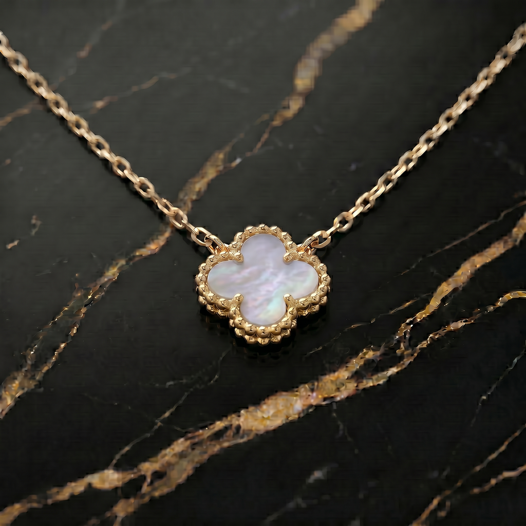 A gold chain necklace with a clover-shaped pendant featuring a white iridescent mother-of-pearl inlay and a textured gold border, displayed on a black marble surface with gold veining.