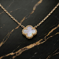 A gold chain necklace with a clover-shaped pendant featuring a white iridescent mother-of-pearl inlay and a textured gold border, displayed on a black marble surface with gold veining.