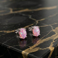 A pair of oval stud earrings with pink opal-like stones in silver-toned settings, displayed on a black marble slab with gold veining.