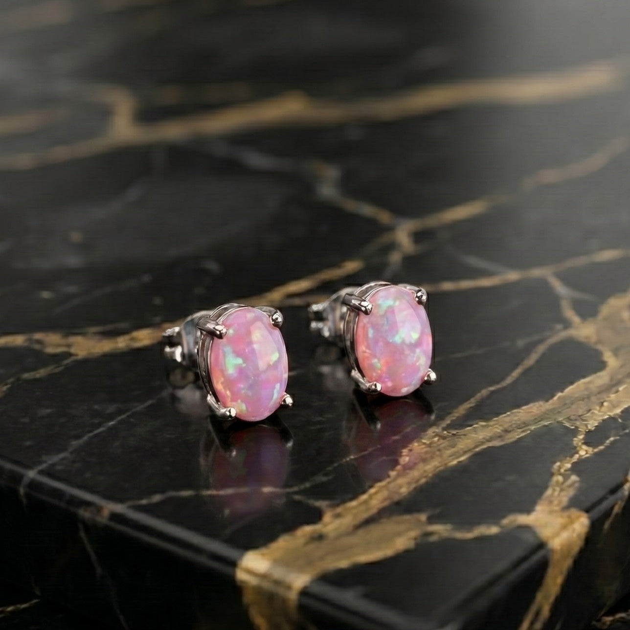 A pair of oval stud earrings with pink opal-like stones in silver-toned settings, displayed on a black marble slab with gold veining.