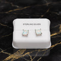 A pair of sterling silver stud earrings with square iridescent opal-like centers, displayed on a white jewelry box labeled 'STERLING SILVER' on a black marble surface with gold veining.
