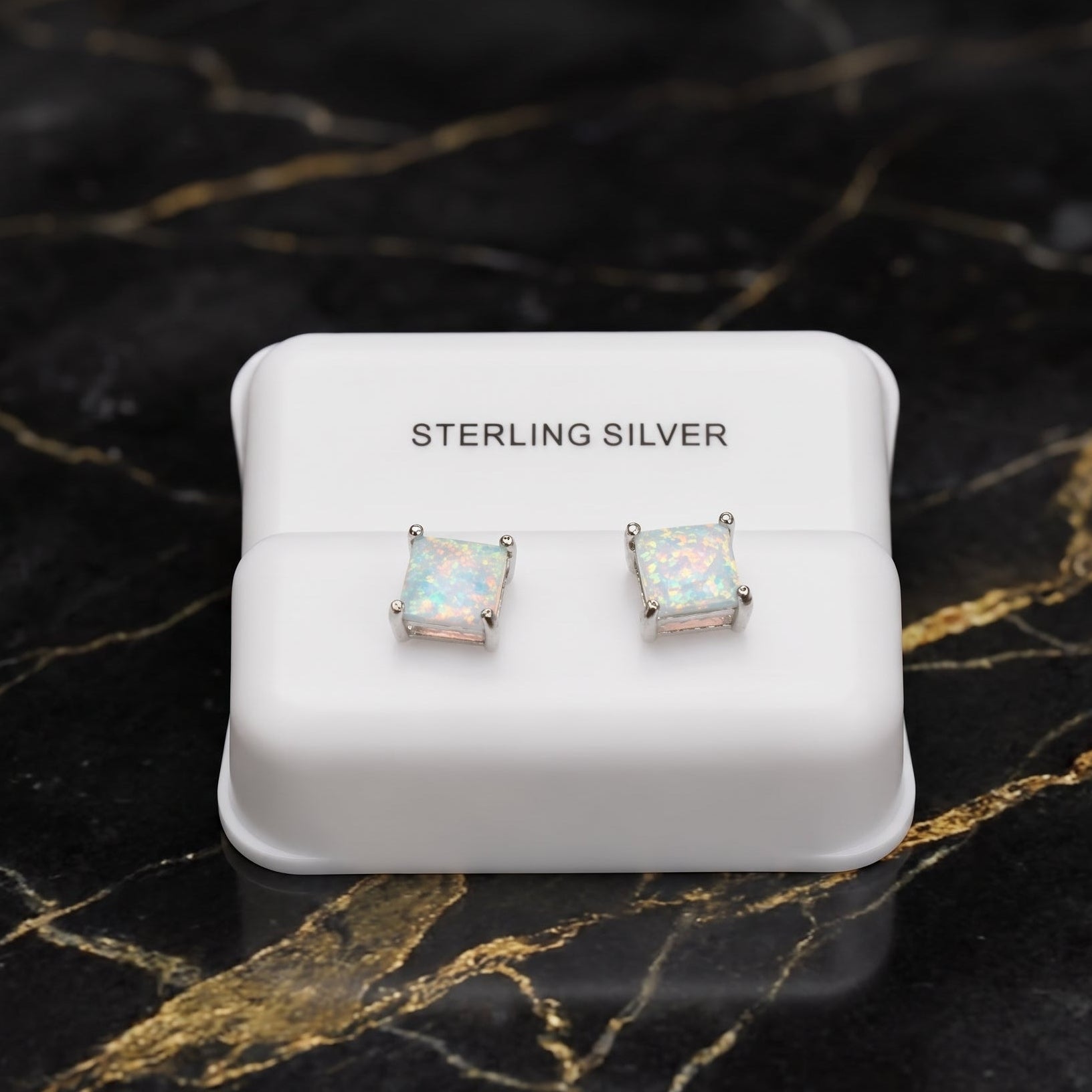 A pair of sterling silver stud earrings with square iridescent opal-like centers, displayed on a white jewelry box labeled 'STERLING SILVER' on a black marble surface with gold veining.