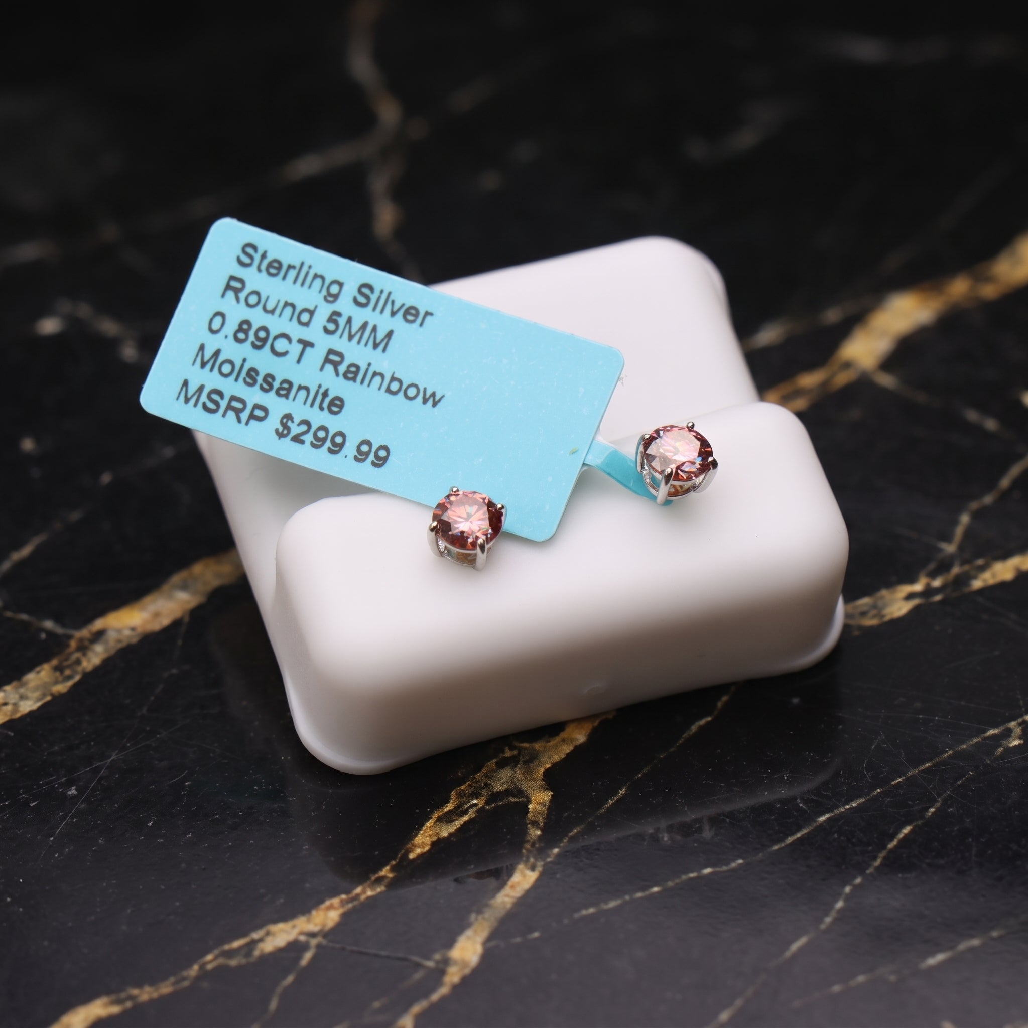 A pair of round-cut pink gemstone stud earrings in sterling silver settings, displayed on a white jewelry box with a blue price tag on a black marble surface.