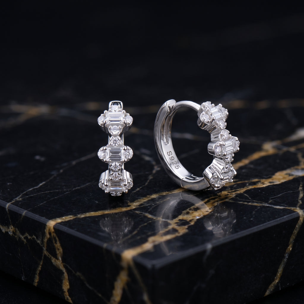 A pair of silver huggie hoop earrings with clear baguette and round-cut stones, displayed on a polished black marble slab with gold veining.