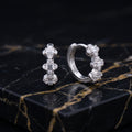 A pair of silver huggie hoop earrings with clear baguette and round-cut stones, displayed on a polished black marble slab with gold veining.