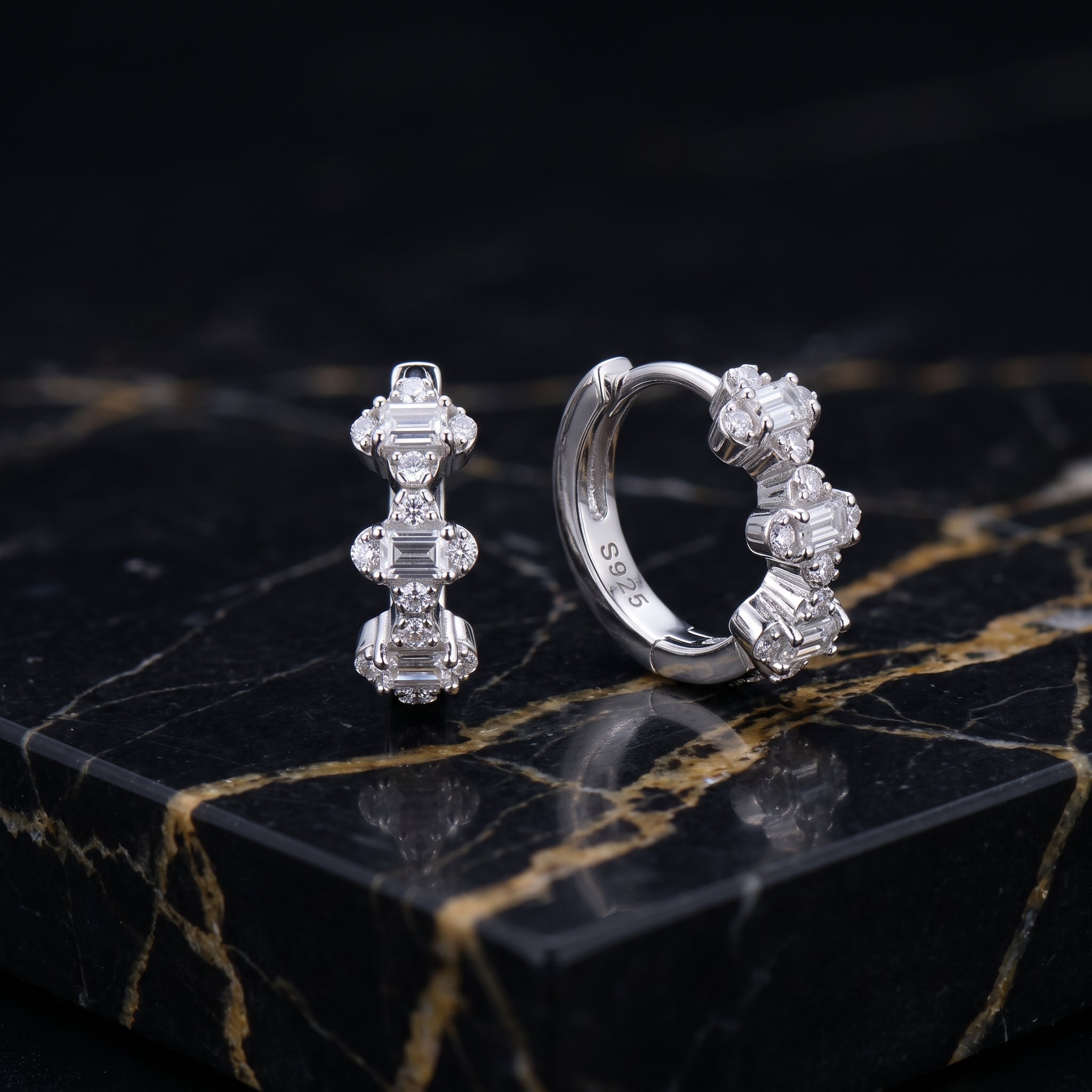 A pair of silver huggie hoop earrings with clear baguette and round-cut stones, displayed on a polished black marble slab with gold veining.