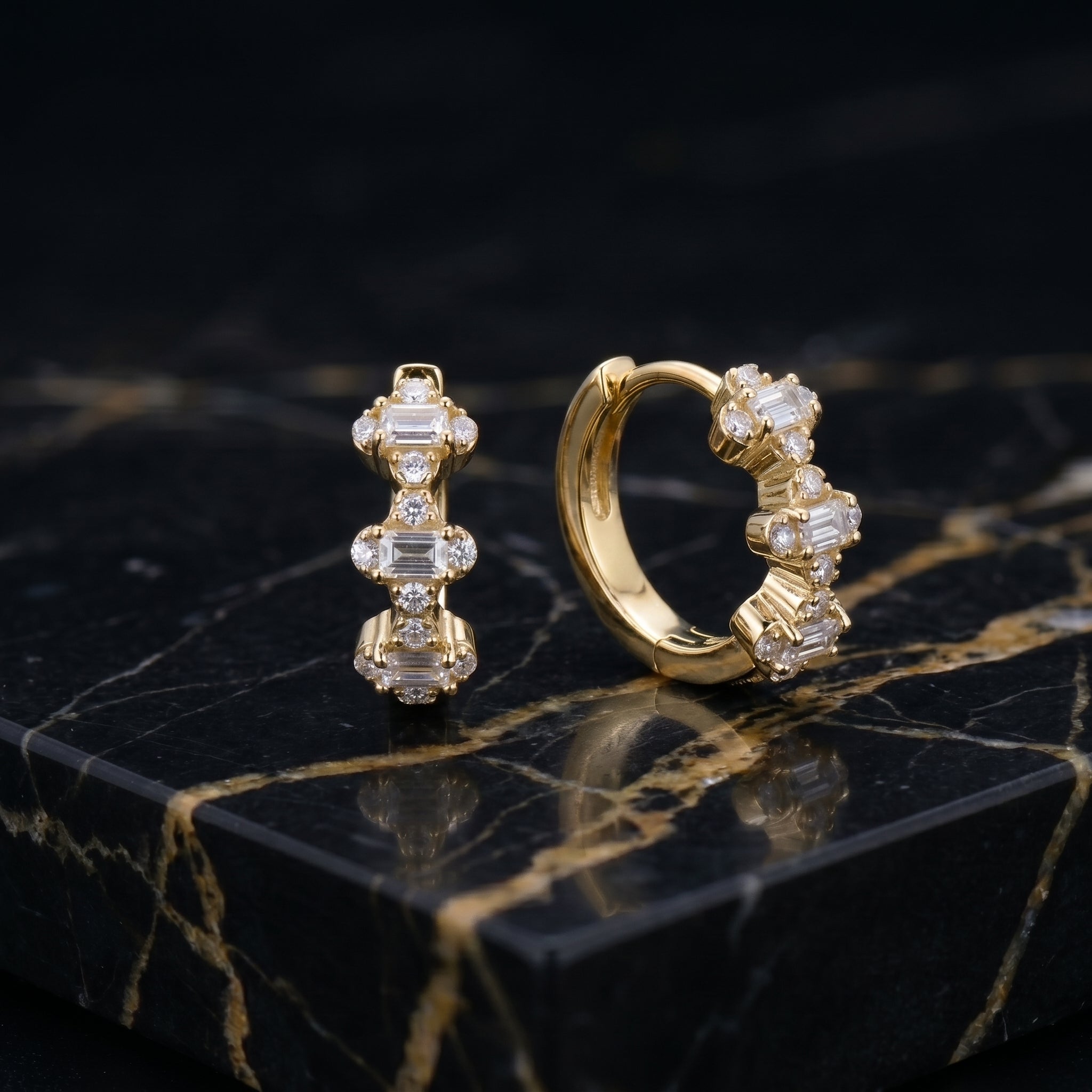 A pair of gold huggie hoop earrings adorned with clear baguette and round crystals, displayed on a black marble surface with gold veining.