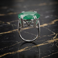 A silver ring featuring a large, faceted green emerald-cut gemstone, displayed on a polished black marble surface with gold veining.