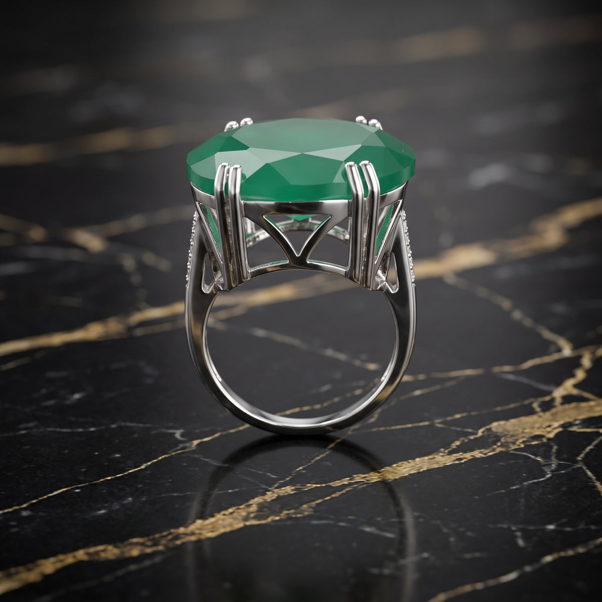 A silver ring featuring a large, faceted green emerald-cut gemstone, displayed on a polished black marble surface with gold veining.