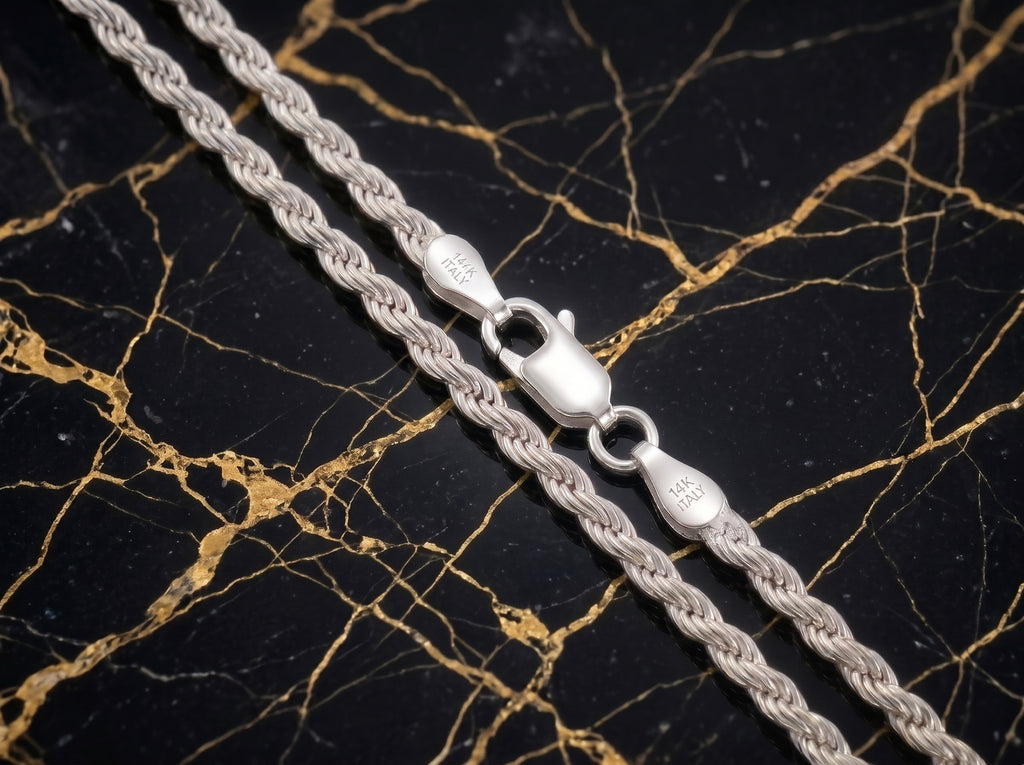 A close-up of a silver rope chain necklace with a lobster clasp, laid diagonally on a black and gold veined marble surface.