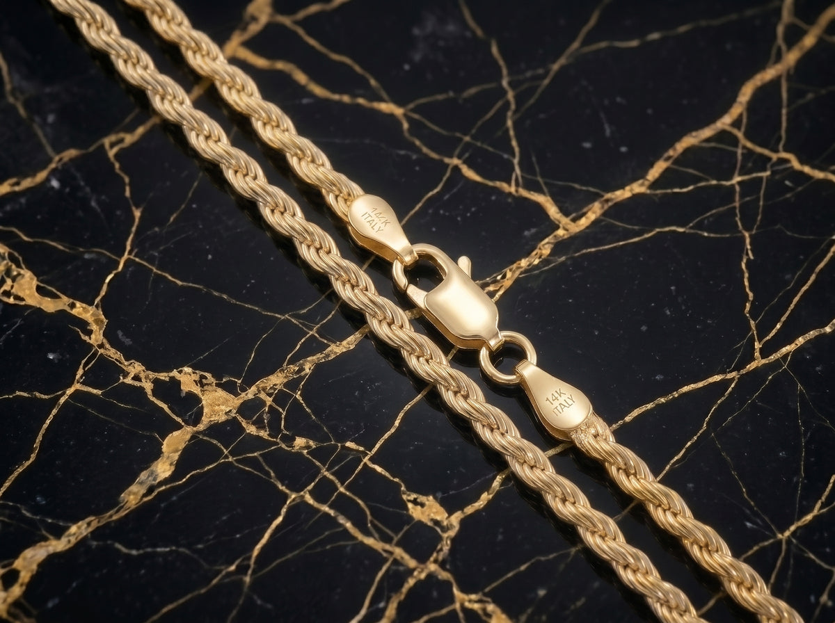 A close-up of a gold rope chain necklace with a lobster clasp, laid diagonally on a black marble background with gold veining.