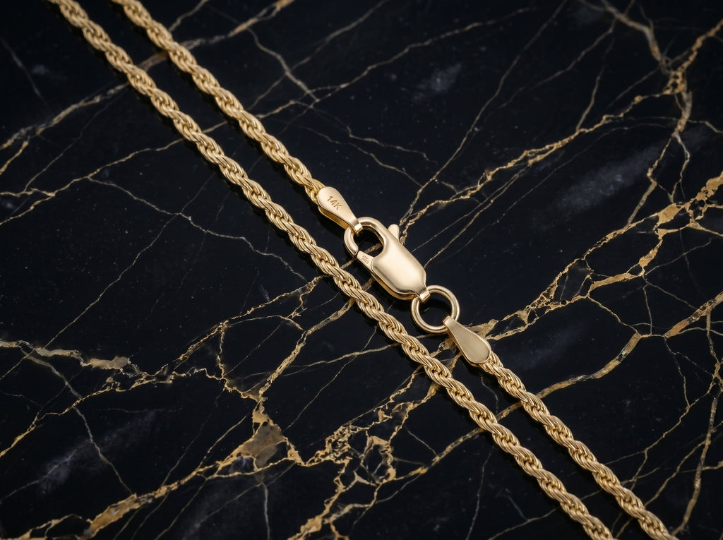 A close-up of a gold rope chain necklace with a lobster clasp, laid diagonally on a black marble surface with gold veining.