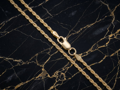 A close-up of a gold rope chain necklace with a lobster clasp, laid diagonally on a black marble surface with gold veining.