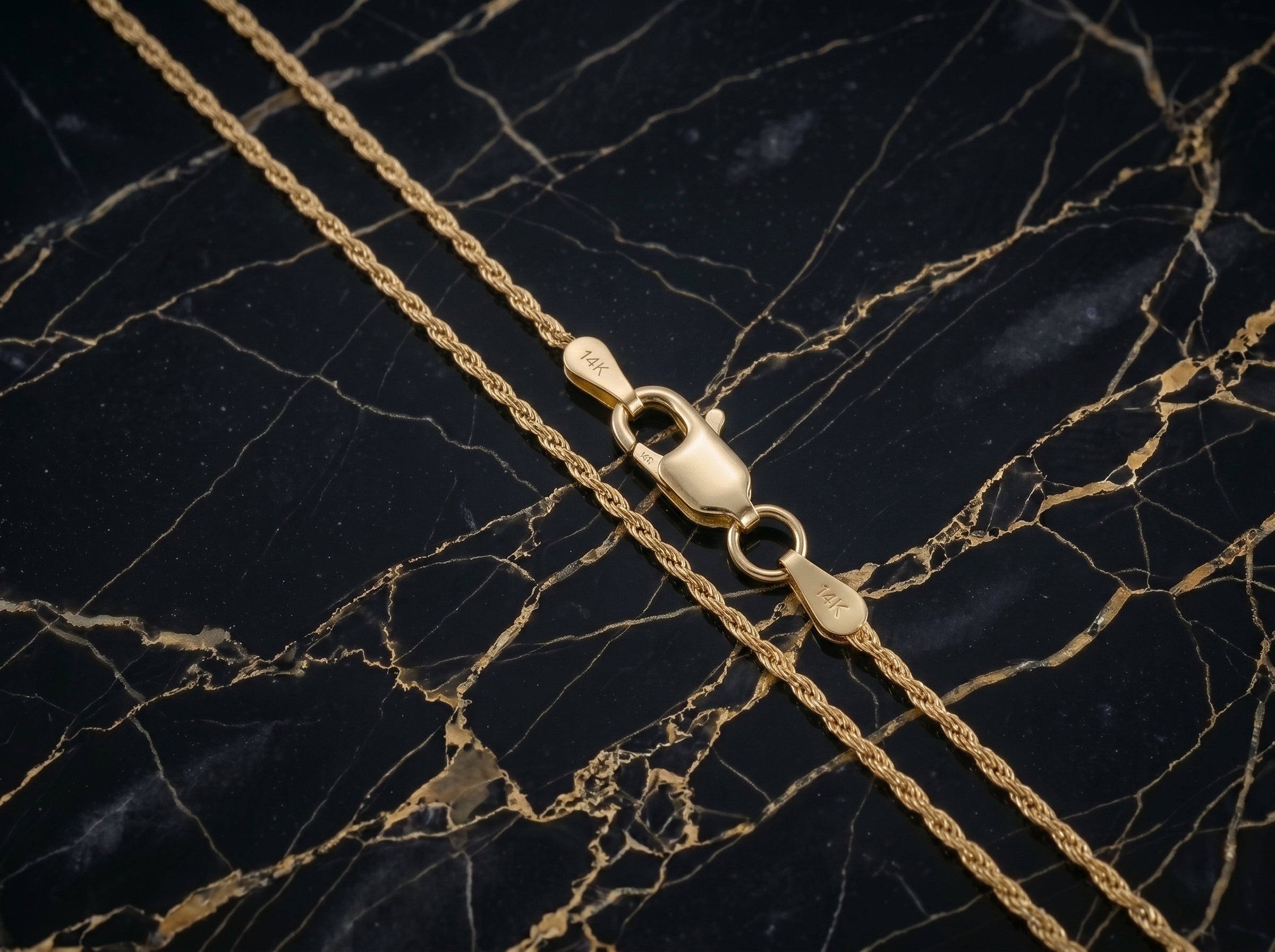 A close-up of a gold chain necklace with a lobster clasp, laid diagonally on a black marble surface with gold veining.