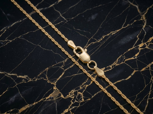 A close-up of a gold chain necklace with a lobster clasp, laid diagonally on a black marble surface with gold veining.