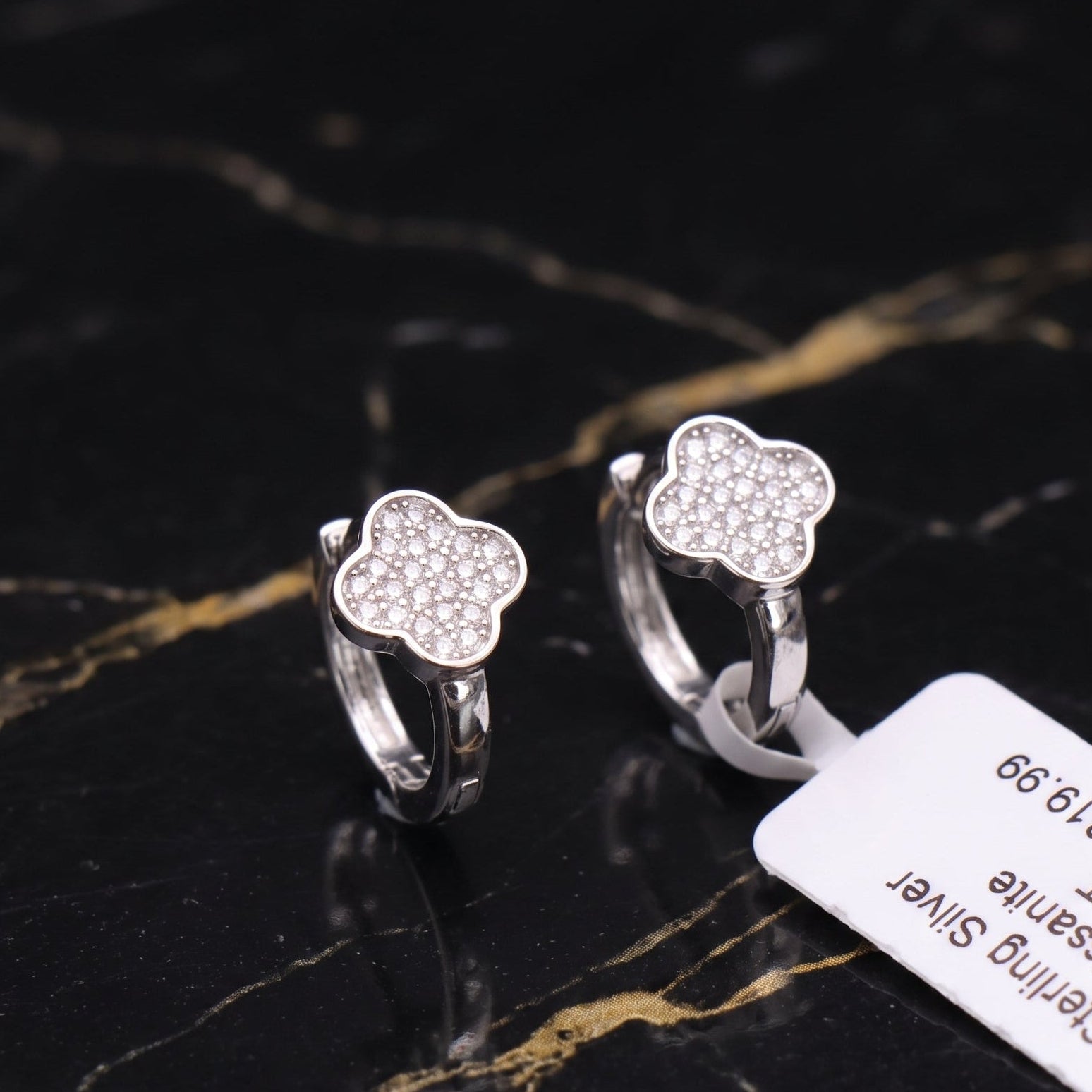 A pair of silver huggie hoop earrings with a clover-shaped face paved with small clear stones, displayed on a black marble surface with a white price tag attached.