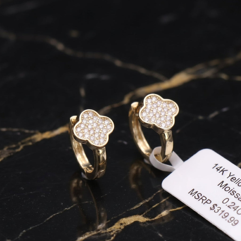 A pair of gold huggie hoop earrings with a four-leaf clover design paved with small clear stones, displayed on a black and gold marble surface next to a white product tag.