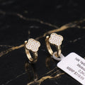 A pair of gold huggie hoop earrings with a four-leaf clover design paved with small clear stones, displayed on a black and gold marble surface next to a white product tag.