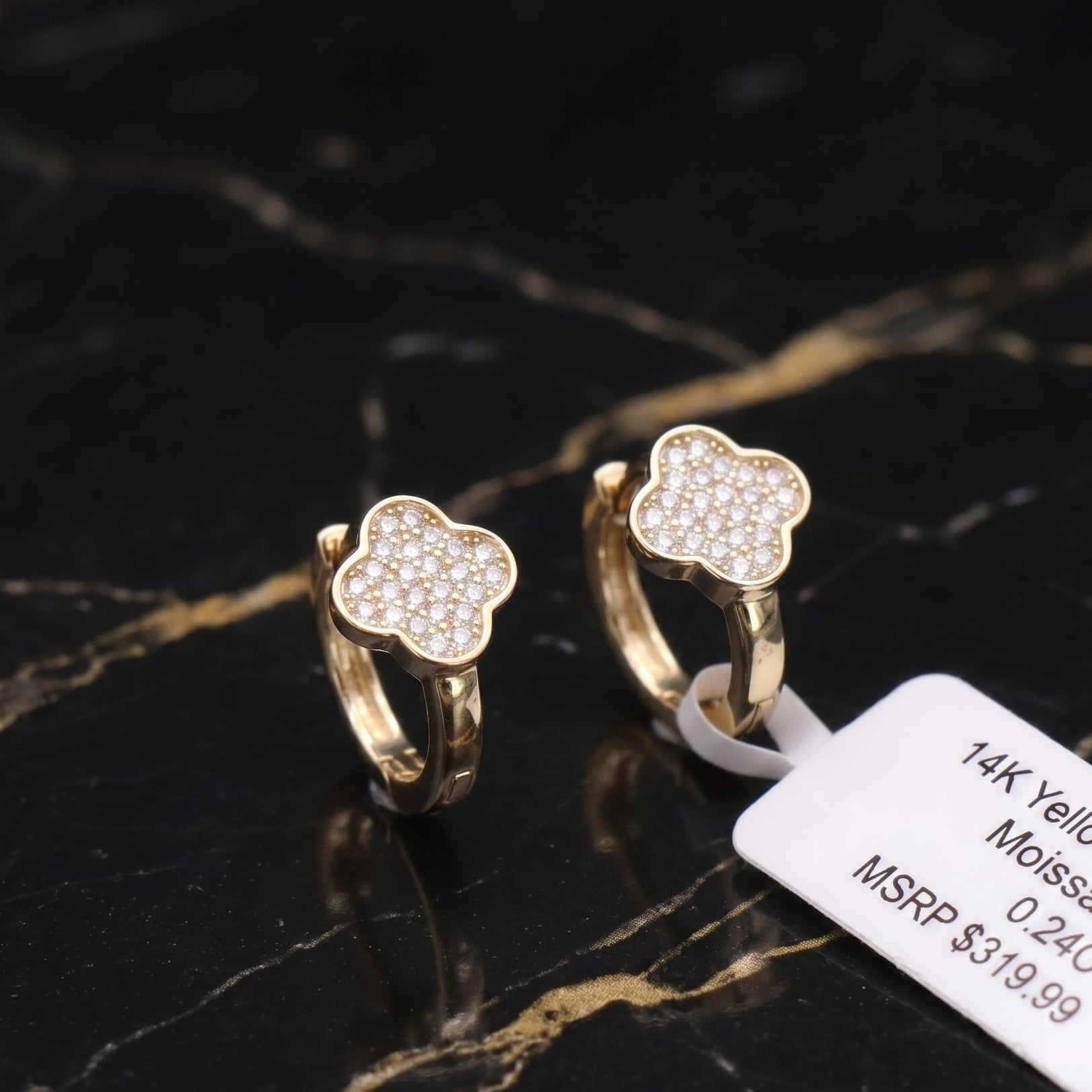A pair of gold huggie hoop earrings with a four-leaf clover design paved with small clear stones, displayed on a black and gold marble surface next to a white product tag.