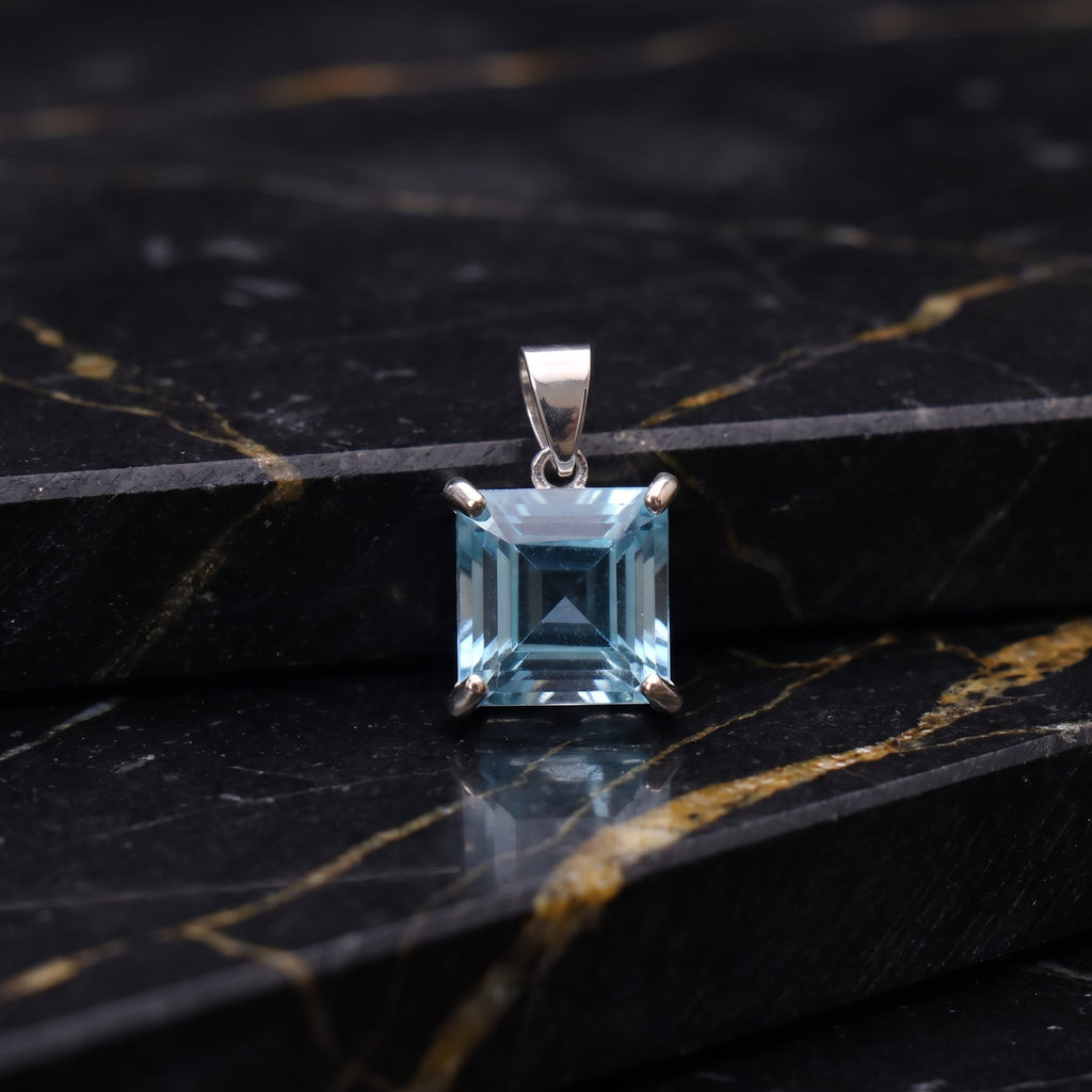 A square-cut light blue gemstone pendant in a silver-toned setting, displayed on a black marble surface with gold veining.