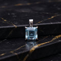 A square-cut light blue gemstone pendant in a silver-toned setting, displayed on a black marble surface with gold veining.