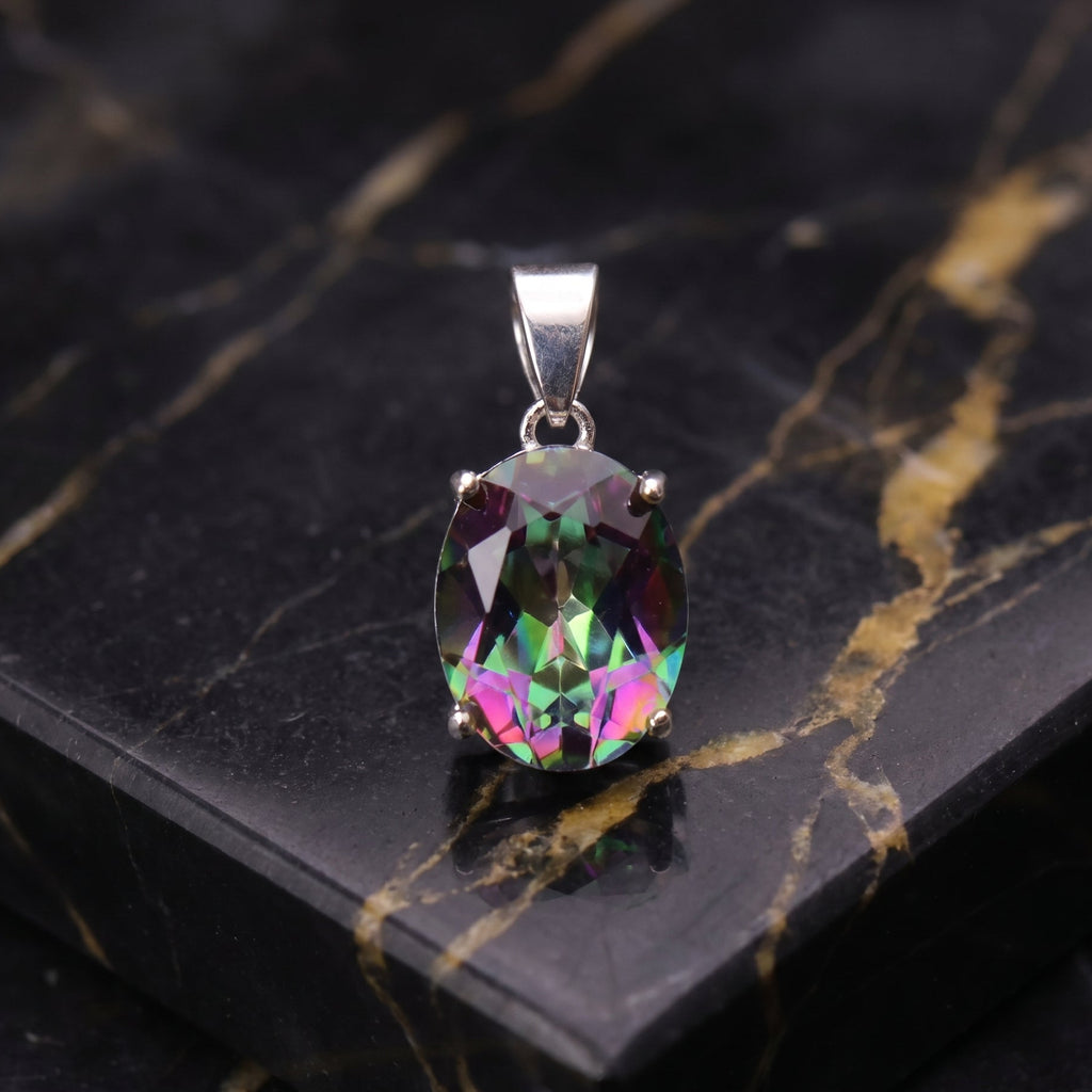 An oval-cut iridescent gemstone pendant in a silver setting, displayed on a black marble slab with gold veining. mystic topaz