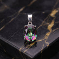 An oval-cut iridescent gemstone pendant in a silver setting, displayed on a black marble slab with gold veining. mystic topaz
