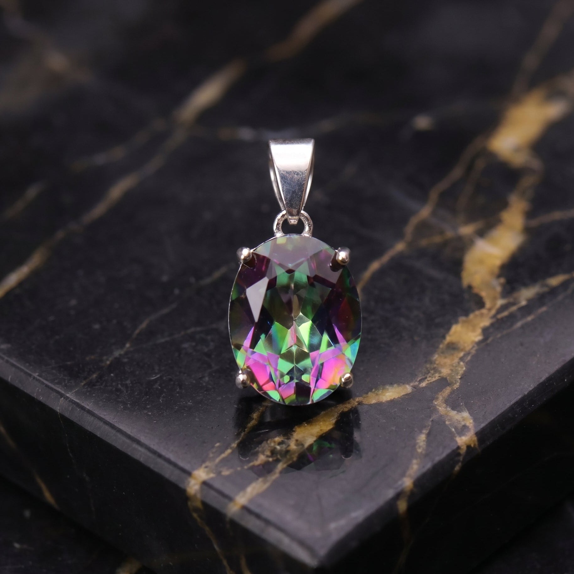 An oval-cut iridescent gemstone pendant in a silver setting, displayed on a black marble slab with gold veining. mystic topaz