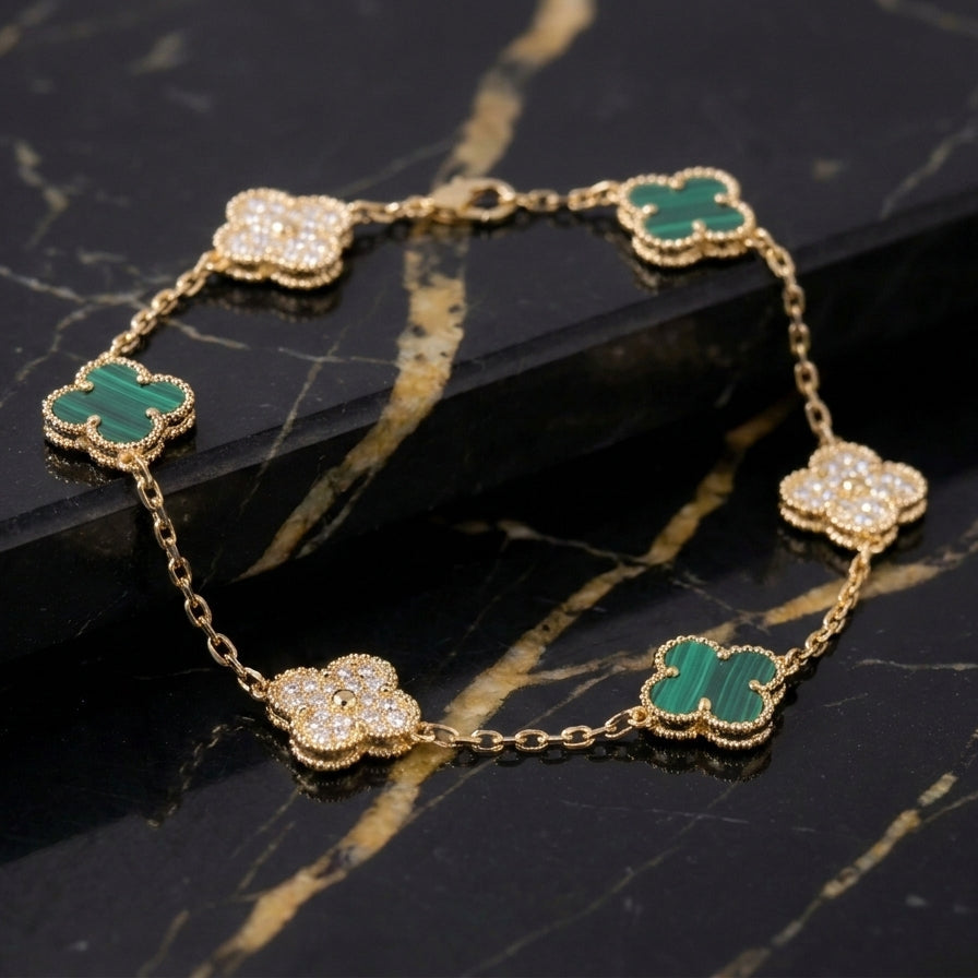A gold chain bracelet featuring five clover-shaped motifs, alternating between green Malachite  and pavé-set clear stones, displayed on a black and gold marble surface.