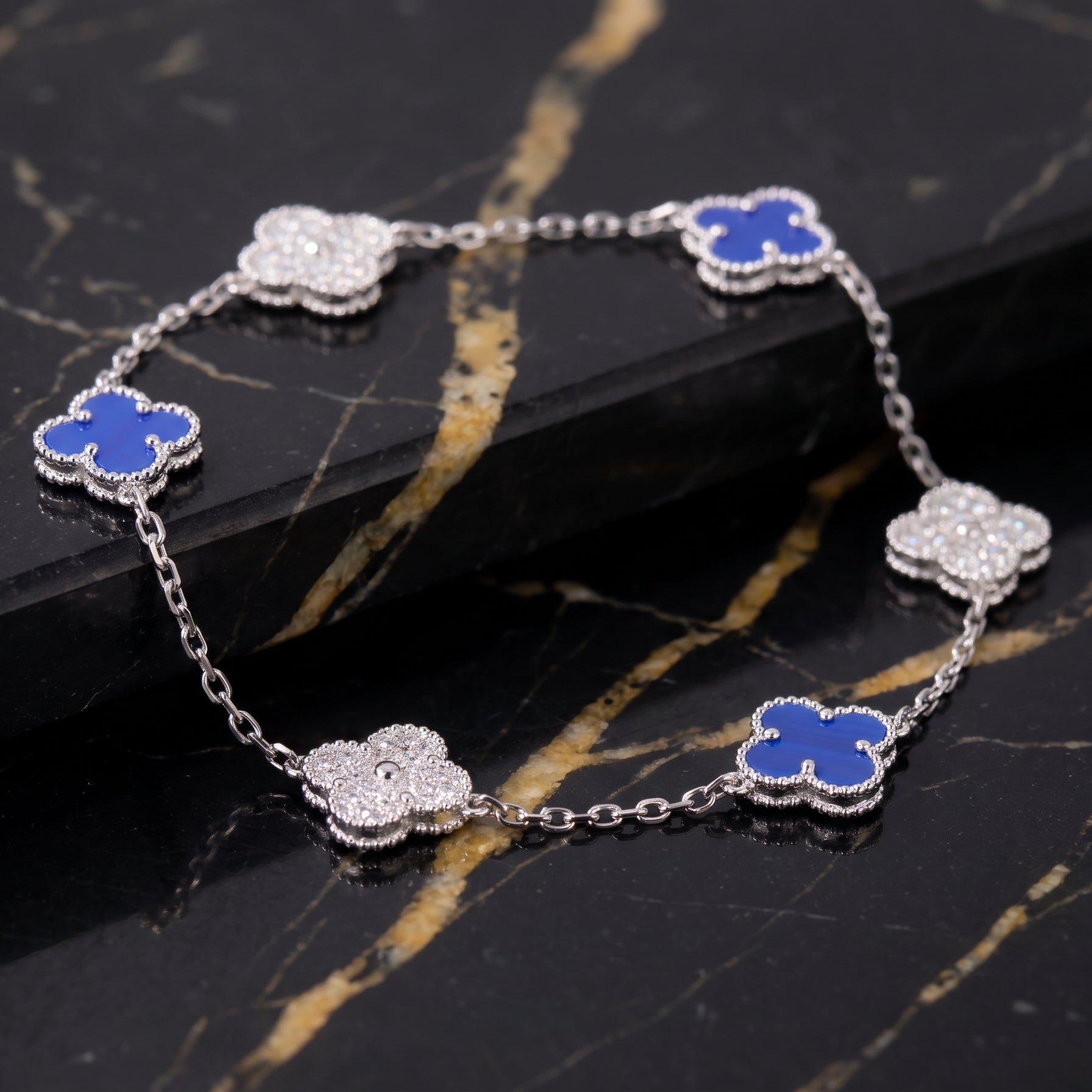A silver-toned chain bracelet with five clover-shaped links, three inlaid with vibrant blue  agate and two with clear pavé stones, displayed on a black and gold marble surface.