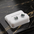 A pair of square stud earrings with iridescent green and purple abalone shell inlays set in silver, displayed on a white plastic jewelry box on a black marble surface.