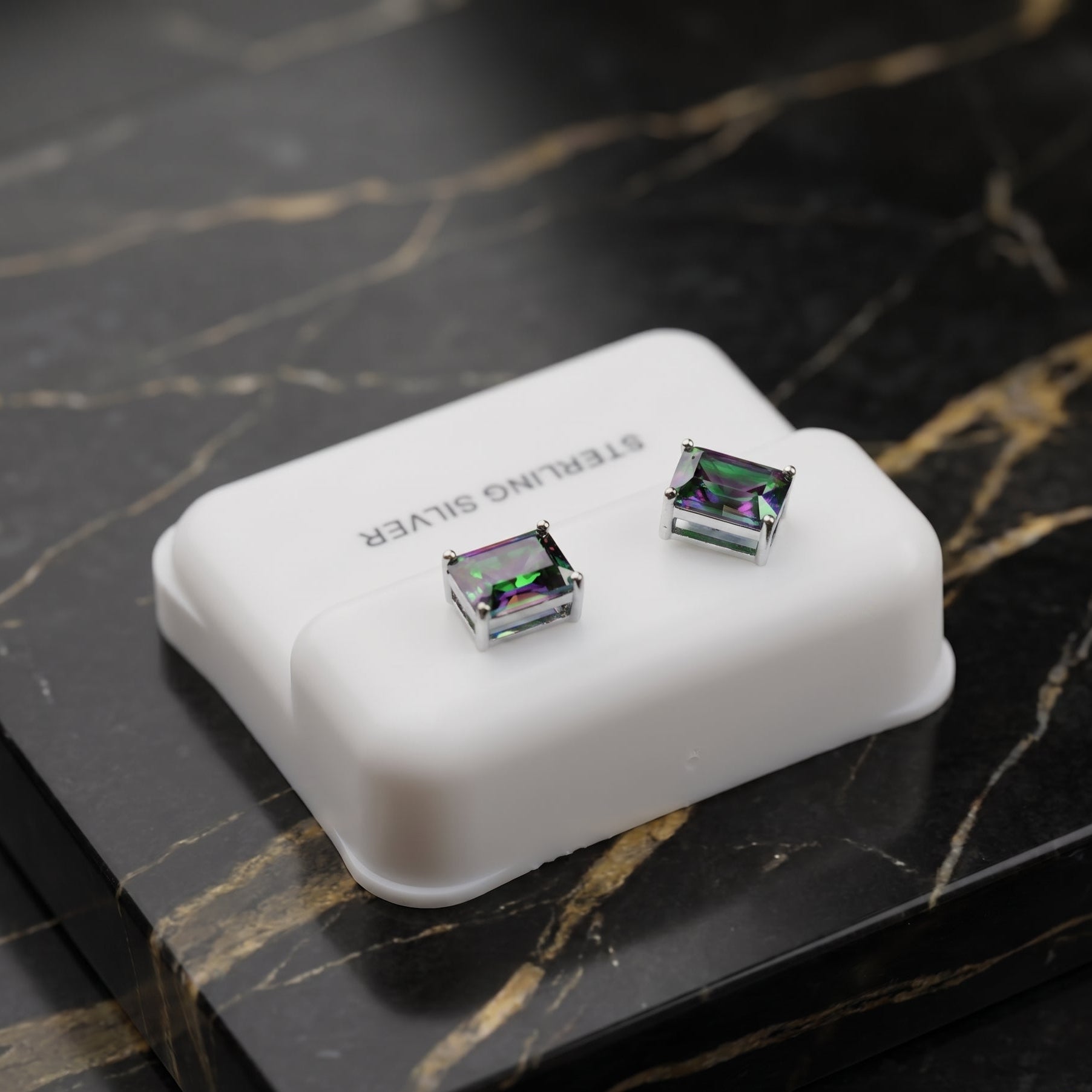 A pair of square stud earrings with iridescent green and purple abalone shell inlays set in silver, displayed on a white plastic jewelry box on a black marble surface.