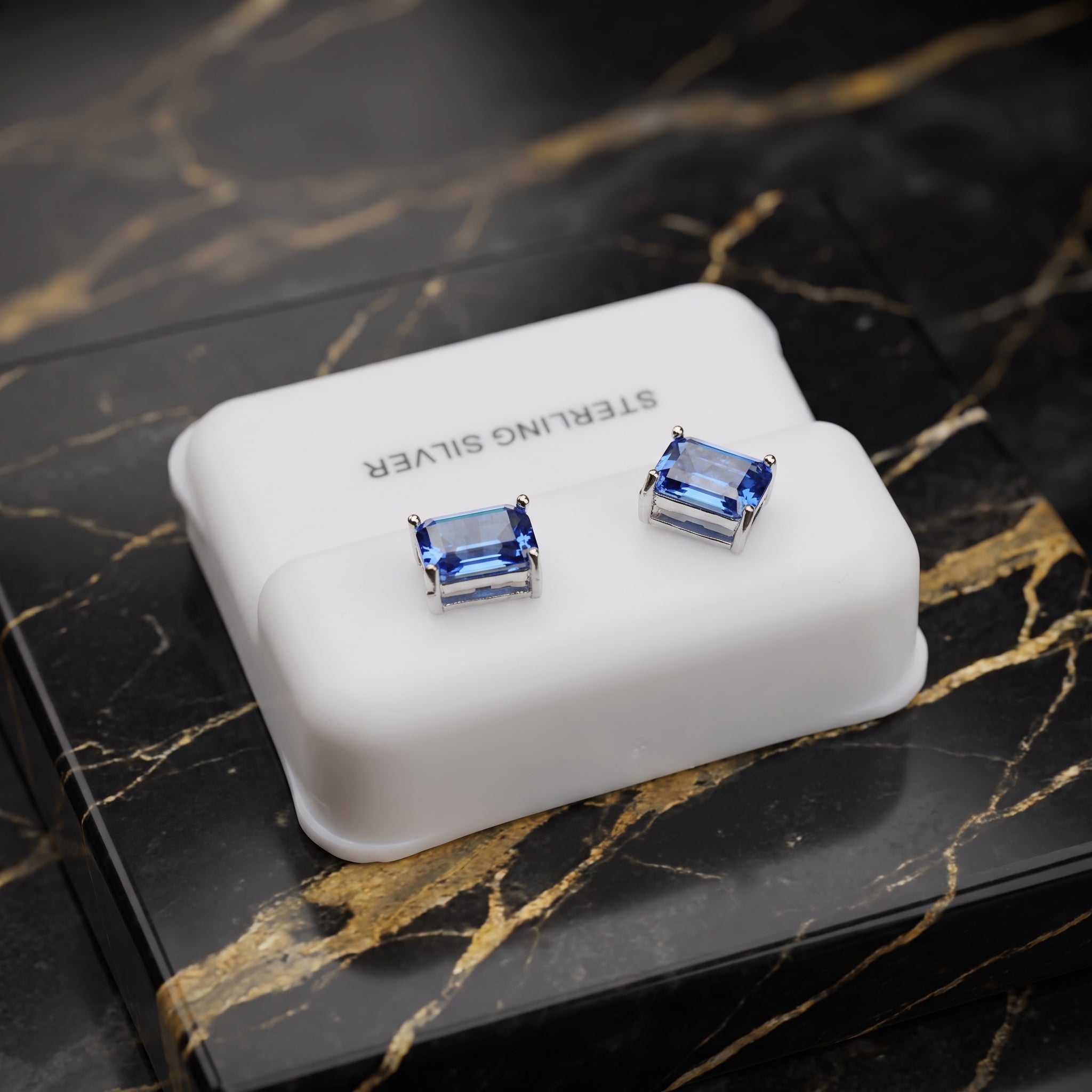 A pair of rectangular blue gemstone stud earrings in silver settings, displayed on a white jewelry box labeled 'STERLING SILVER' on a black marble surface.