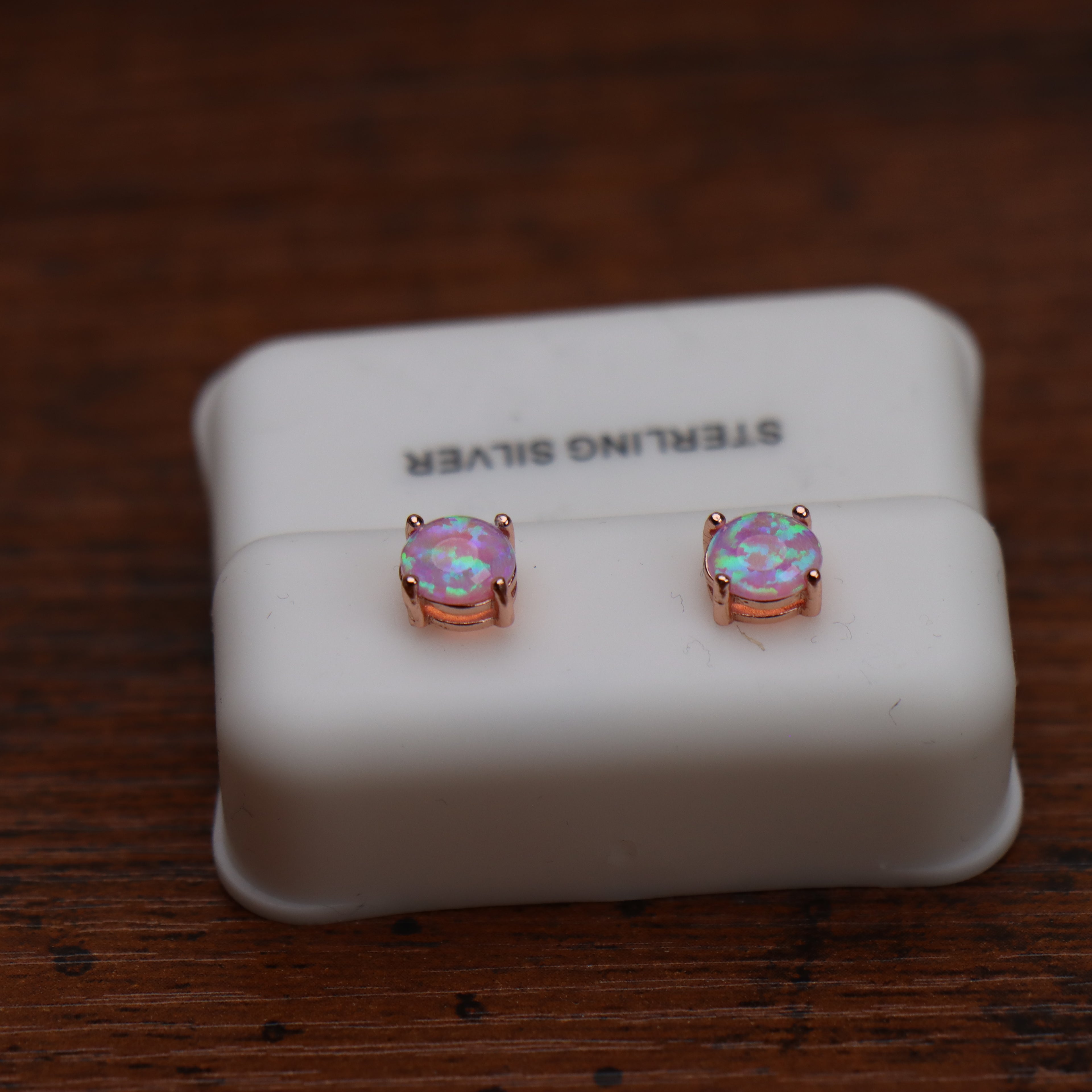 A pair of round bezel-set opal stud earrings in rose gold, displayed on a white jewelry display block that reads 'STERLING SILVER', set on a dark wooden surface.