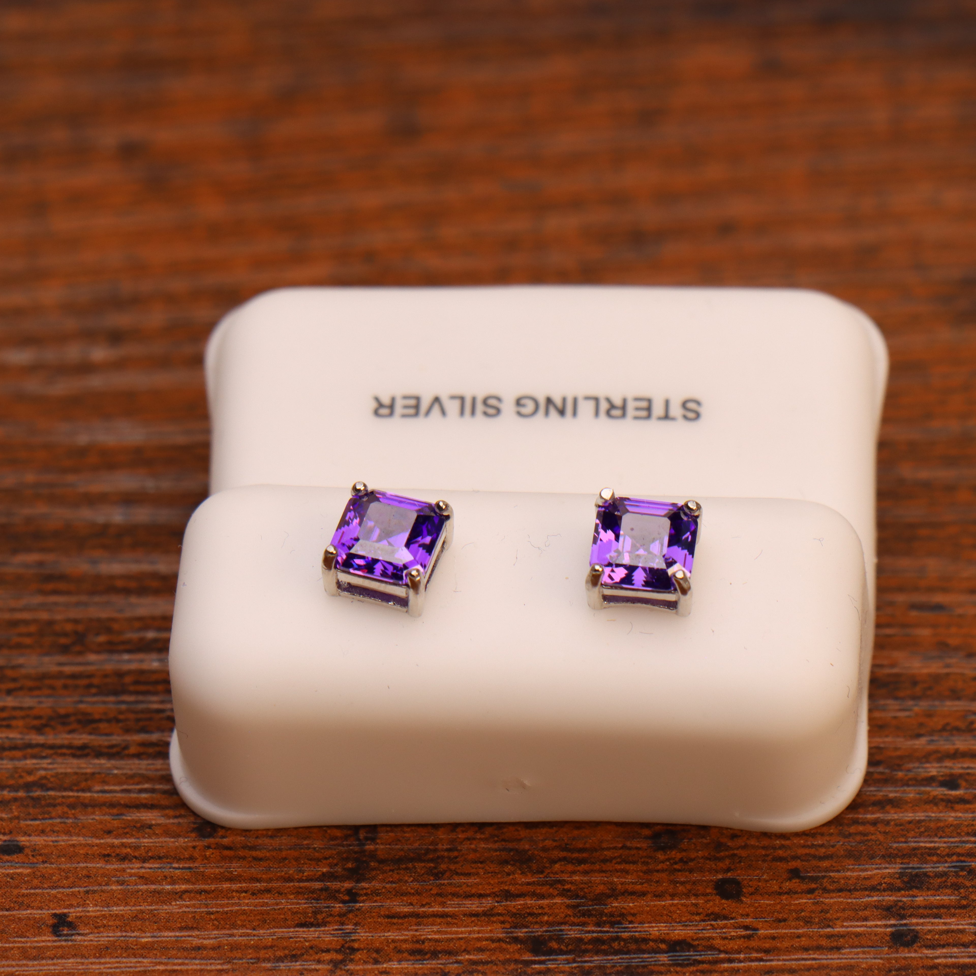 Purple CZ Earrings, Square Asscher Cut in 925 Sterling Silver, displayed on a white holder atop a wooden surface.