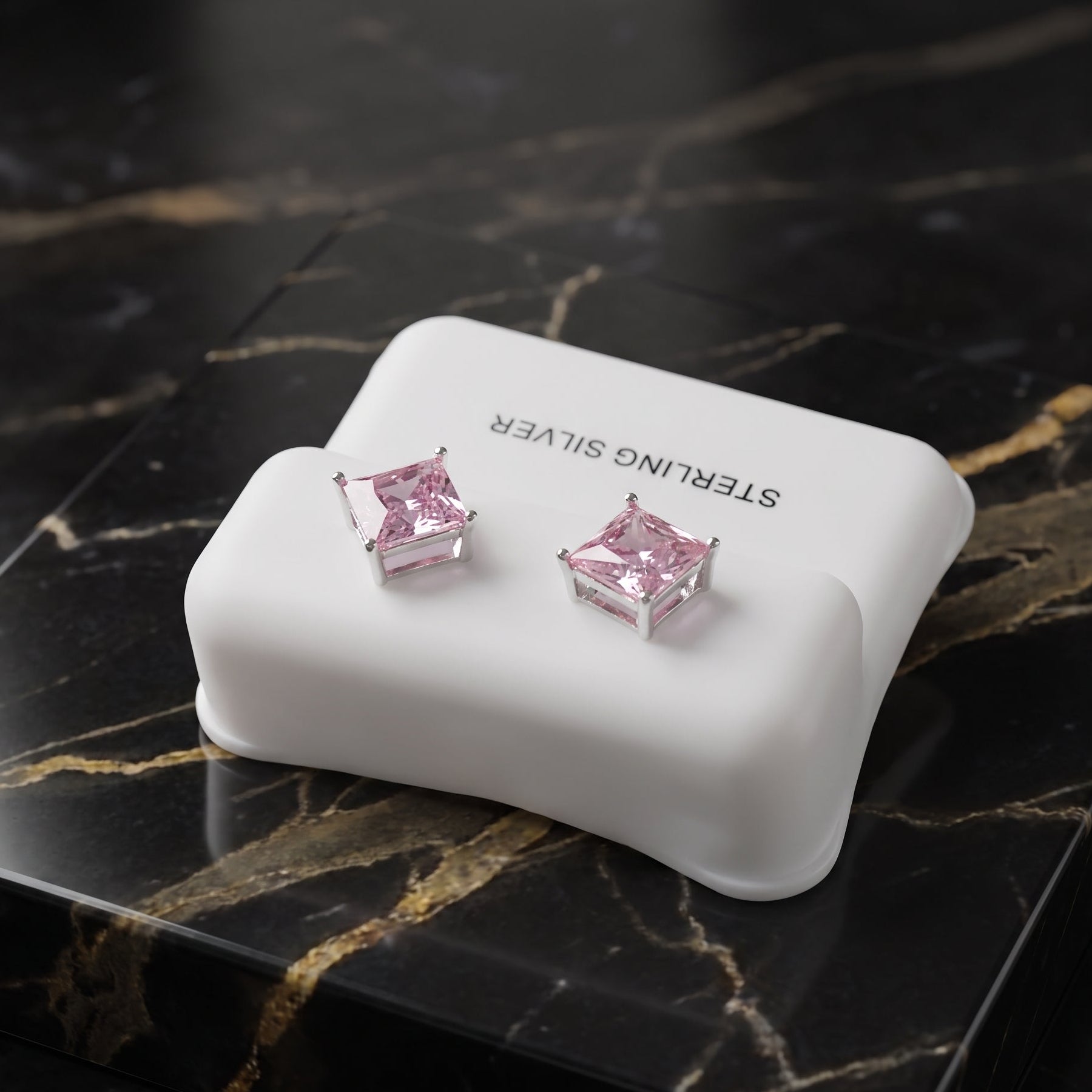 A pair of pink gemstone stud earrings in silver settings, displayed on a white silicone stand labeled 'STERLING SILVER' on a black marble surface.