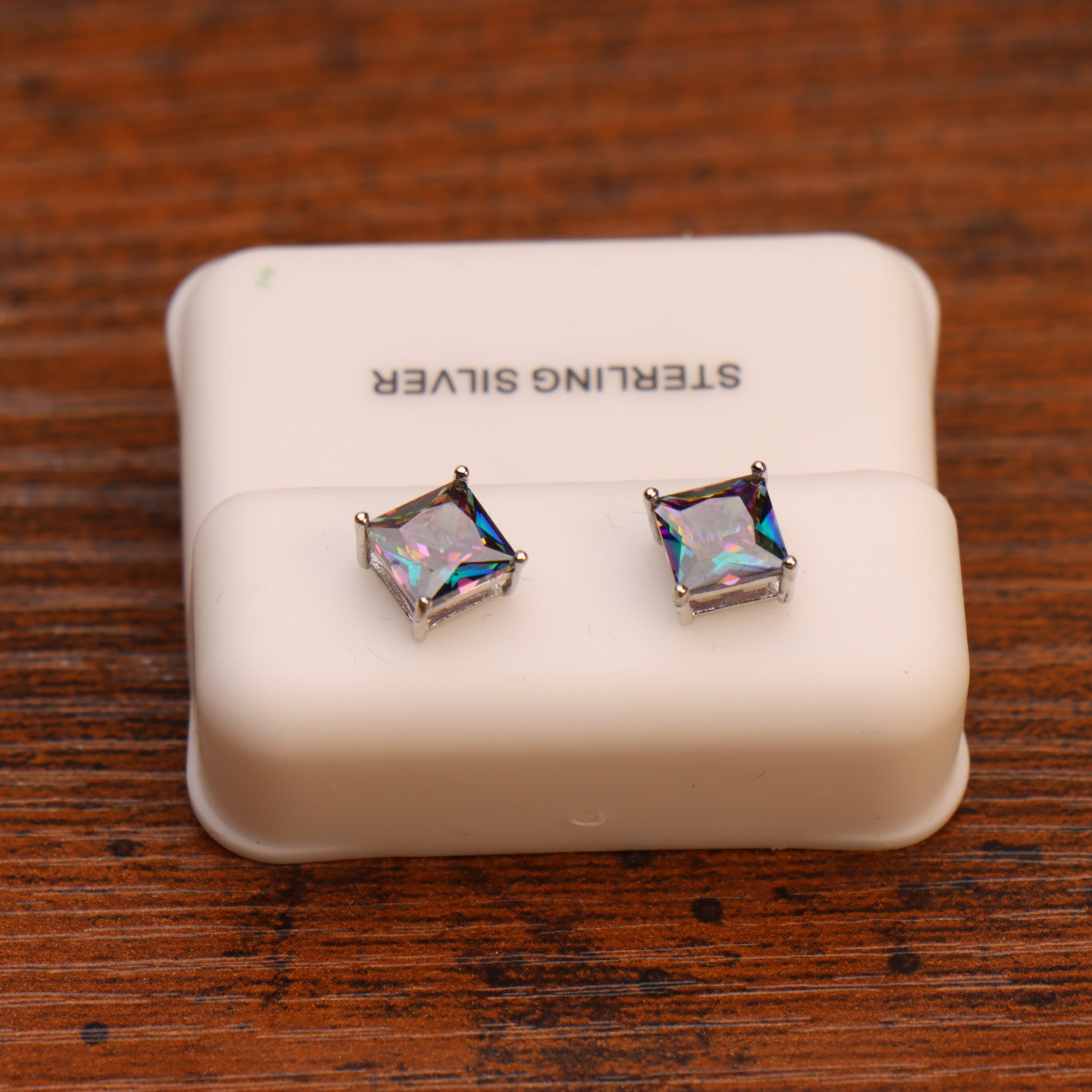 A pair of Mystic Topaz Cz square princess cut earrings in 925 sterling silver, displayed on a white padded holder labeled STERLING SILVER and placed on a wooden surface.
