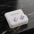A pair of square-cut purple gemstone stud earrings in silver settings, displayed on a white jewelry stand with the text 'STERLING SILVER' on a black marble surface with gold veining.