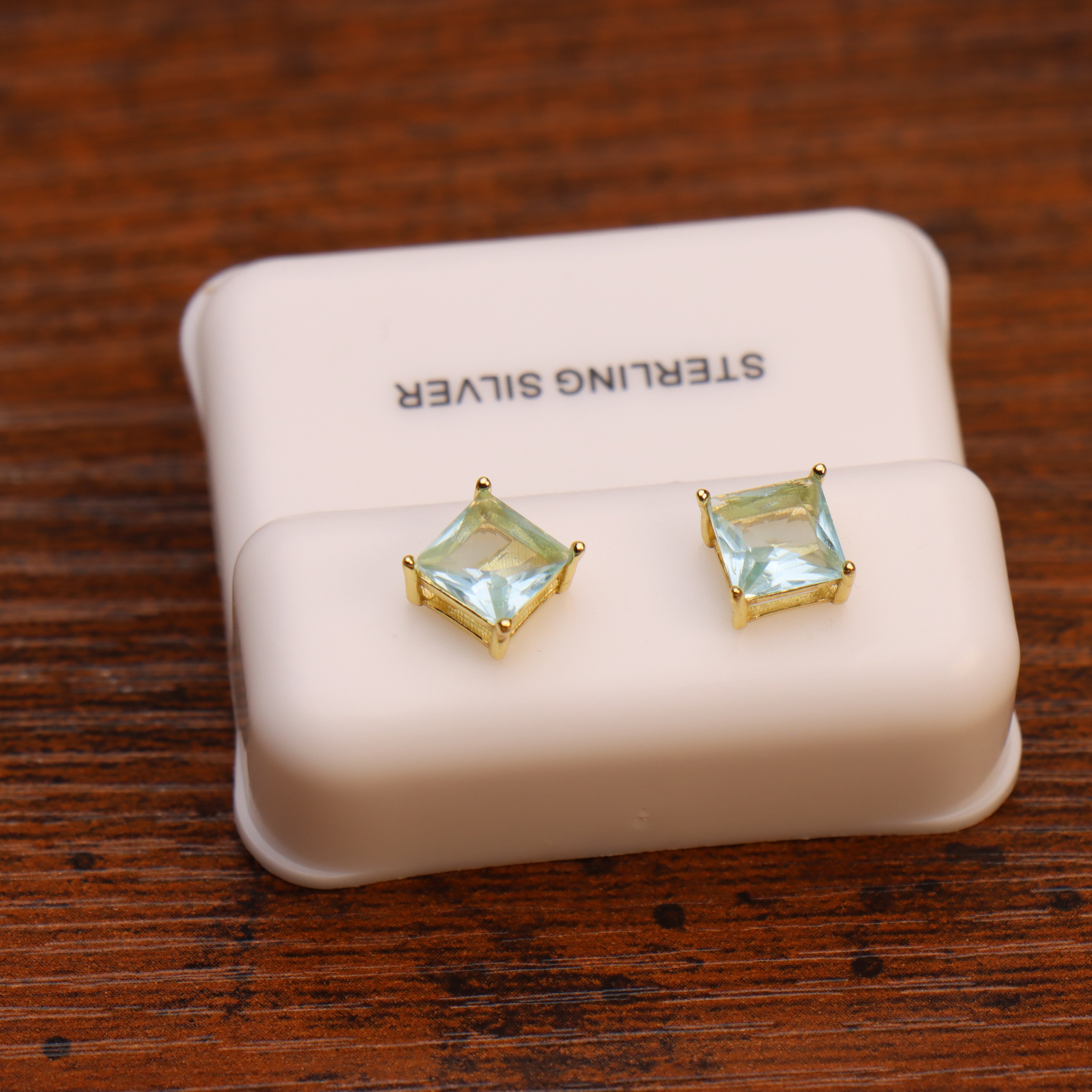A pair of square-cut blue topaz CZ earrings, set in 14k gold over 925 sterling silver, displayed on a white STERLING SILVER box atop a wooden surface.