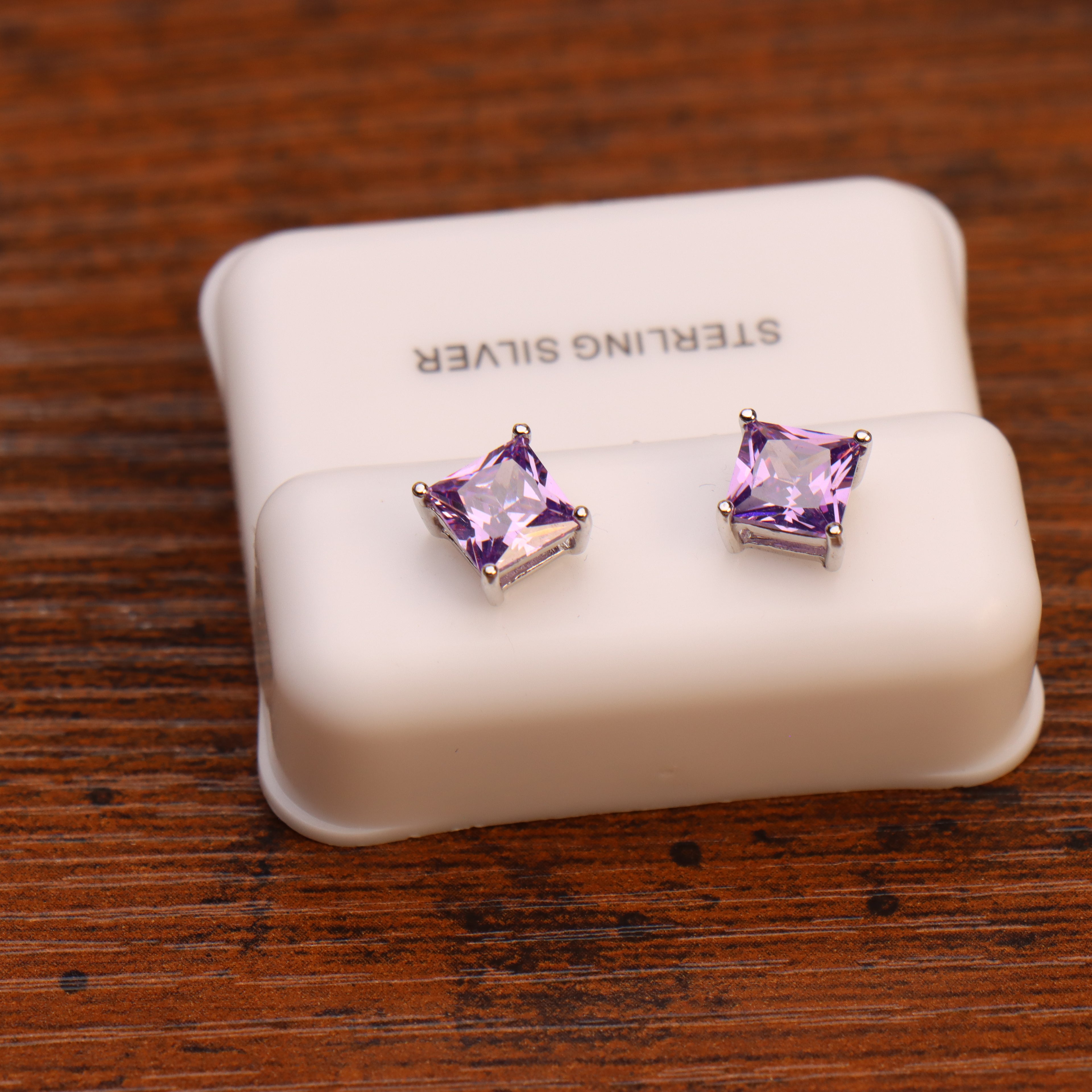 A pair of Amethyst Cz Earrings, square princess cut in 925 sterling silver, displayed on a white case labeled STERLING SILVER and placed on a wooden surface.