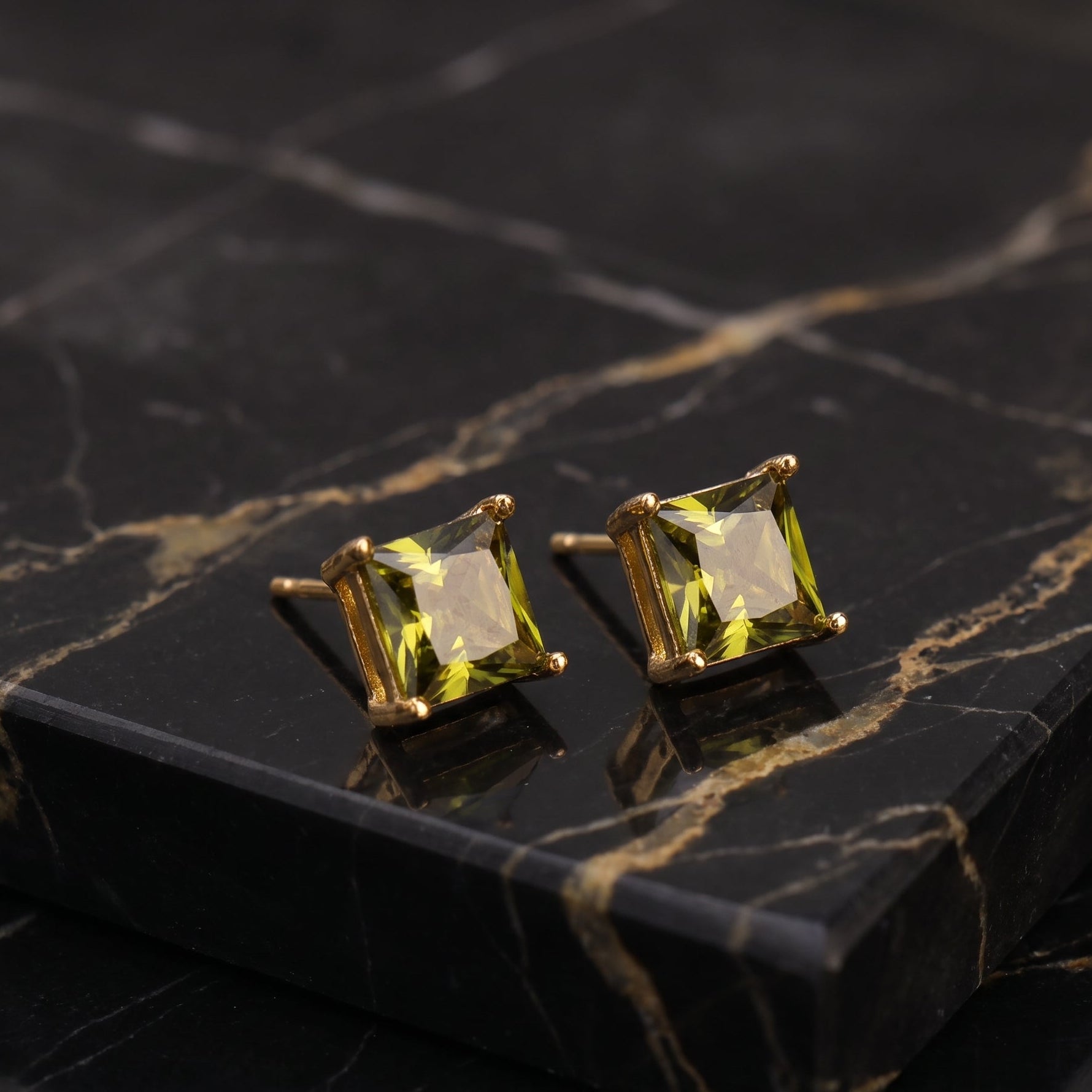 A pair of gold stud earrings with square-cut yellow-green gemstones, displayed on a black marble block with gold veining.