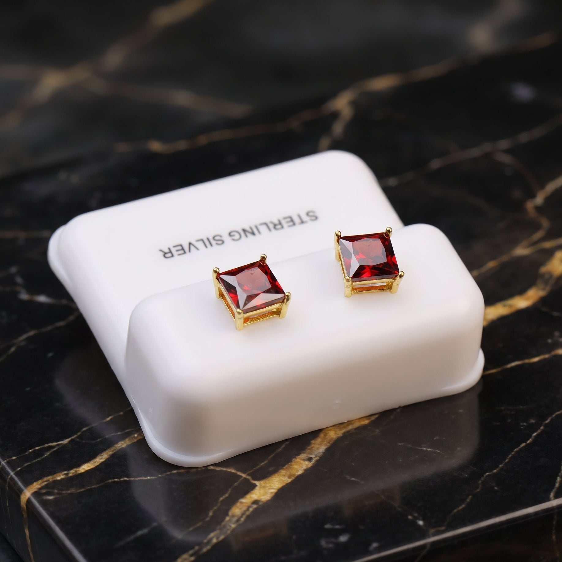 A pair of square stud earrings with deep red gemstones in gold settings, displayed on a white jewelry stand labeled 'STERLING SILVER' on a black marble surface.