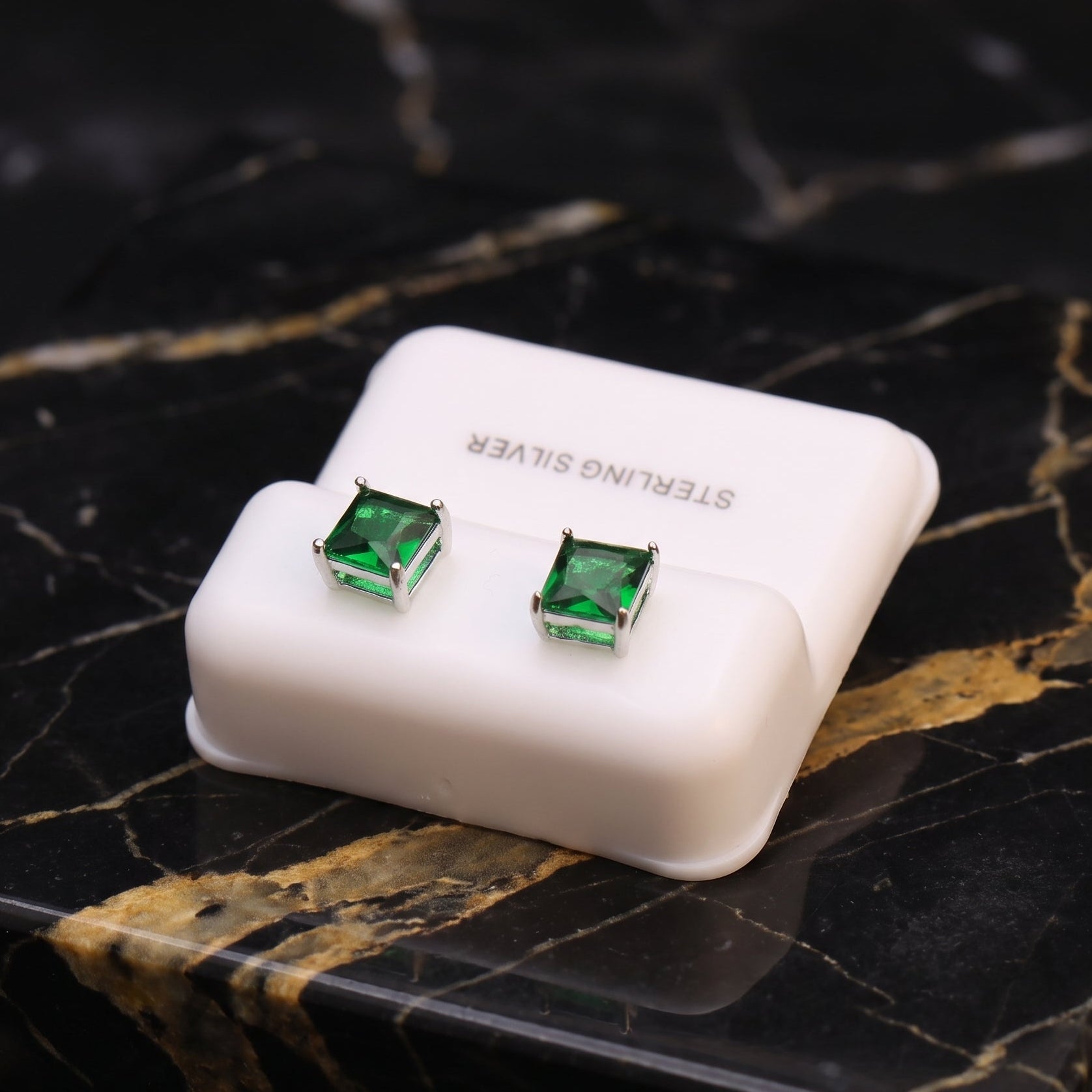A pair of square-cut green stud earrings in silver settings, displayed on a small white jewelry box placed on a black and gold marble surface.