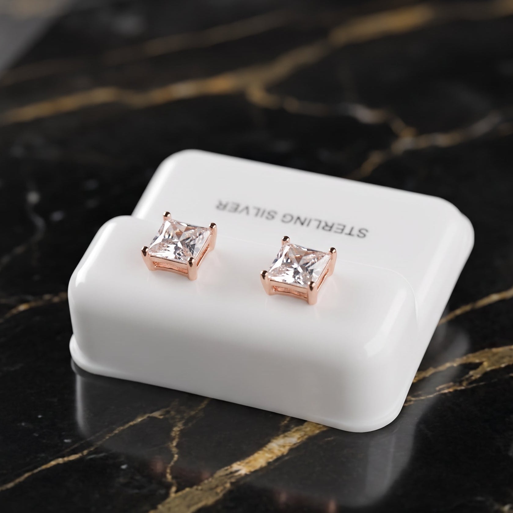 A pair of princess-cut clear cubic zirconia stud earrings in rose gold settings, displayed on a white 'STERLING SILVER' branded stand on a black marble surface with gold veining.