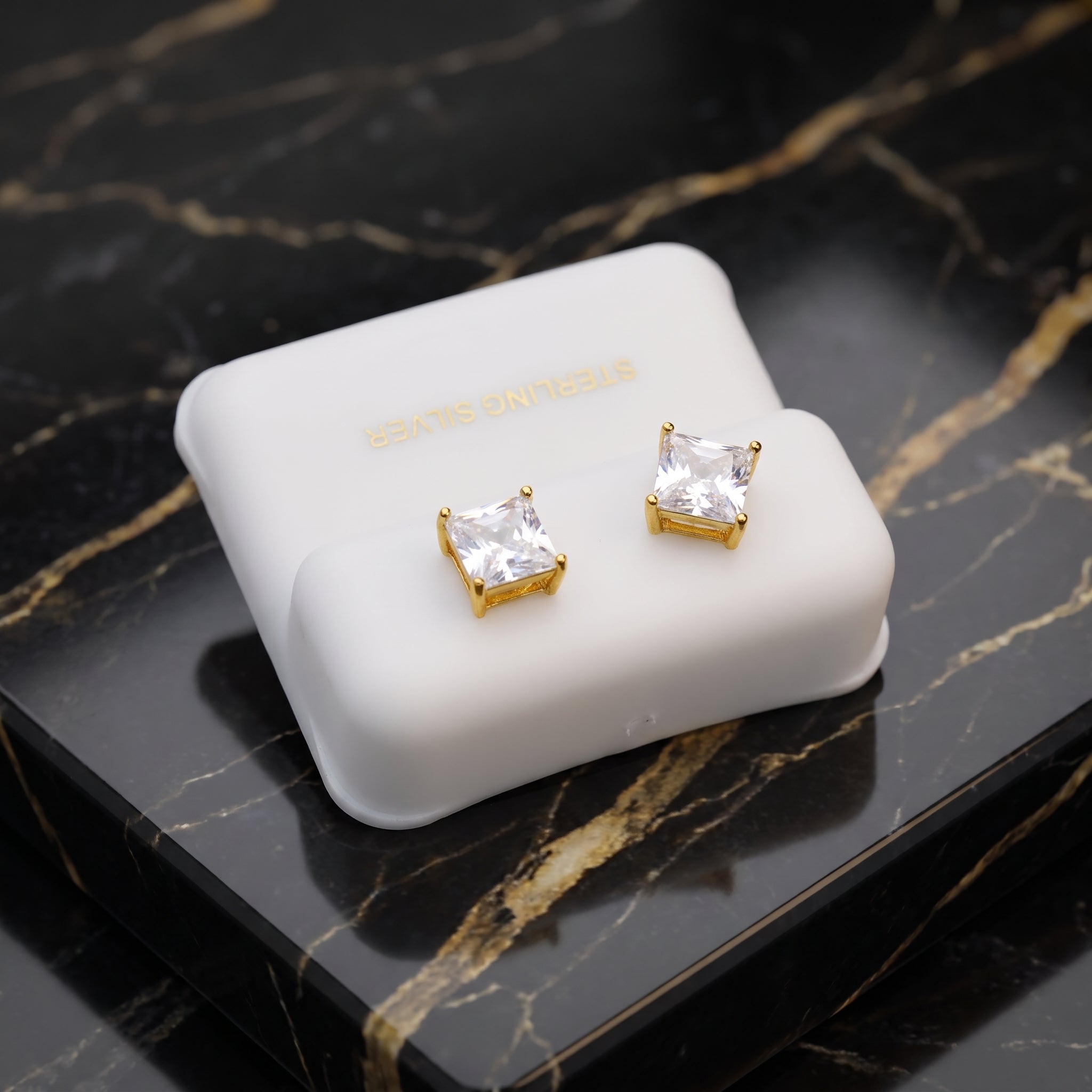 A pair of gold stud earrings with clear square-cut stones, displayed on a white jewelry box set on a black marble surface.