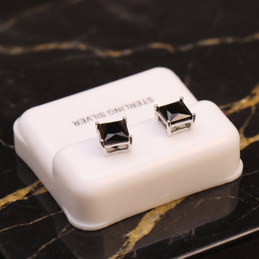 A pair of square-cut black gemstone stud earrings in silver settings, displayed on a small white plastic jewelry box labeled 'STERLING SILVER' on a black marble surface with gold veining.