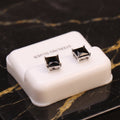 A pair of square-cut black gemstone stud earrings in silver settings, displayed on a small white plastic jewelry box labeled 'STERLING SILVER' on a black marble surface with gold veining.