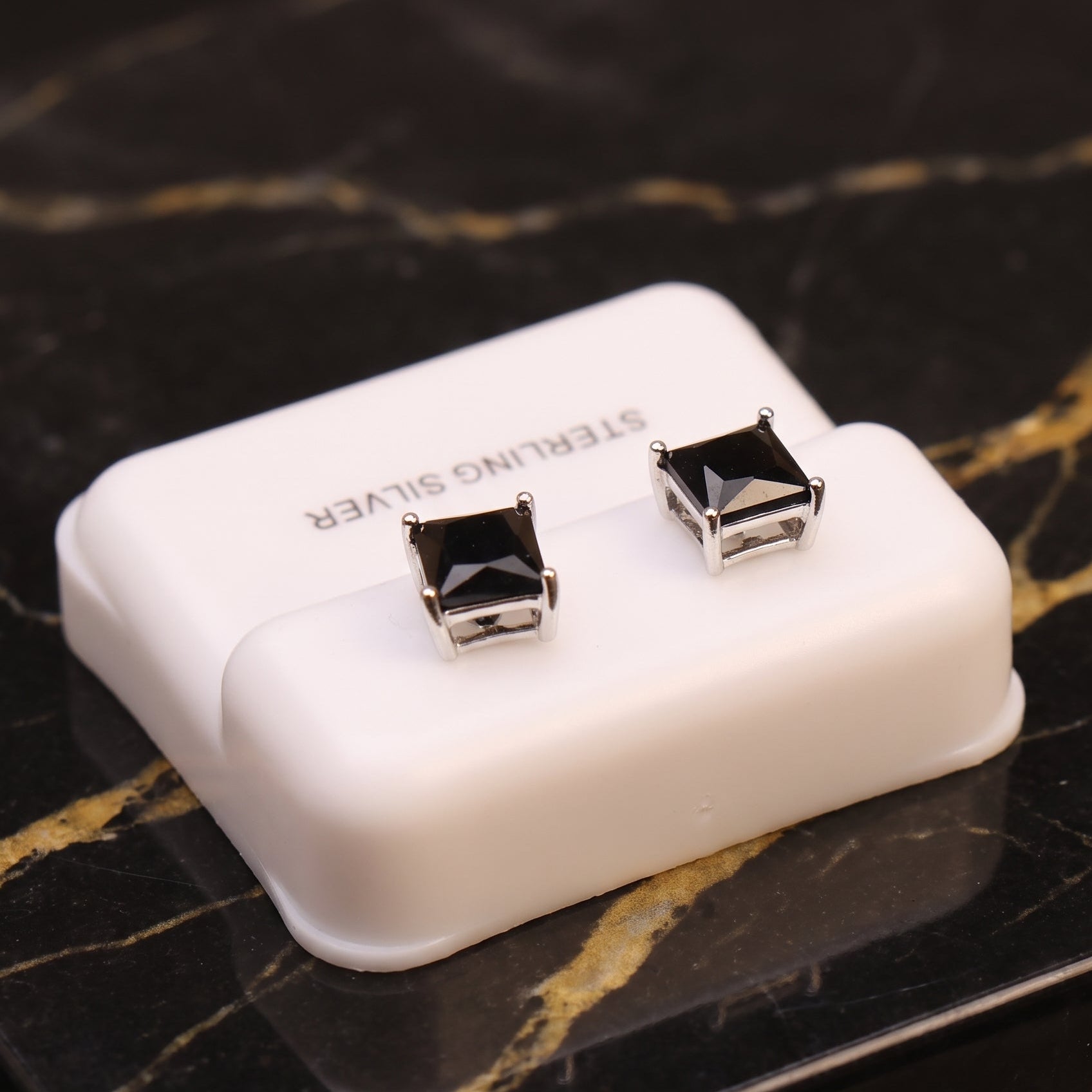 A pair of square-cut black gemstone stud earrings in silver settings, displayed on a small white plastic jewelry box labeled 'STERLING SILVER' on a black marble surface with gold veining.