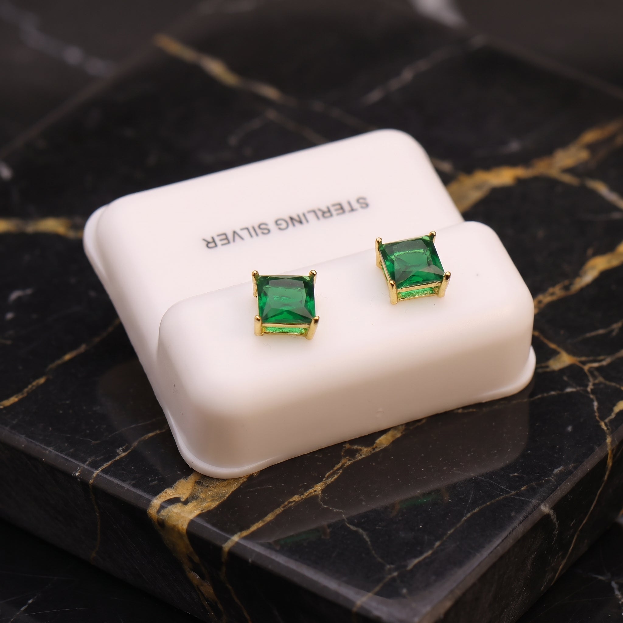 A pair of square-cut green gemstone stud earrings with gold settings, displayed on a white jewelry box labeled 'STERLING SILVER' on a black marble surface.